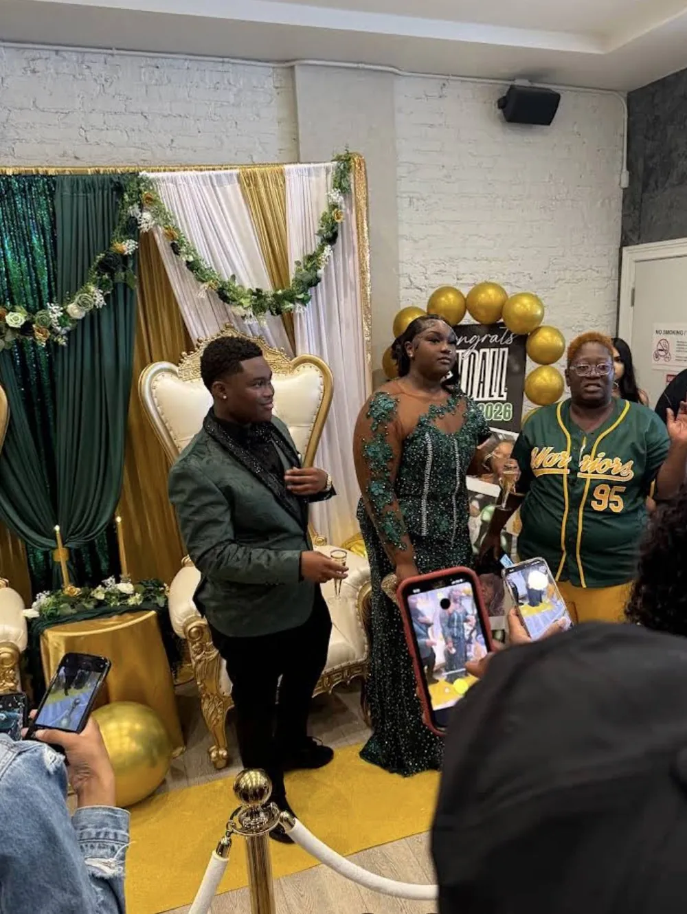 Graduates posing in front of a green and gold backdrop with a throne chair and balloon arch