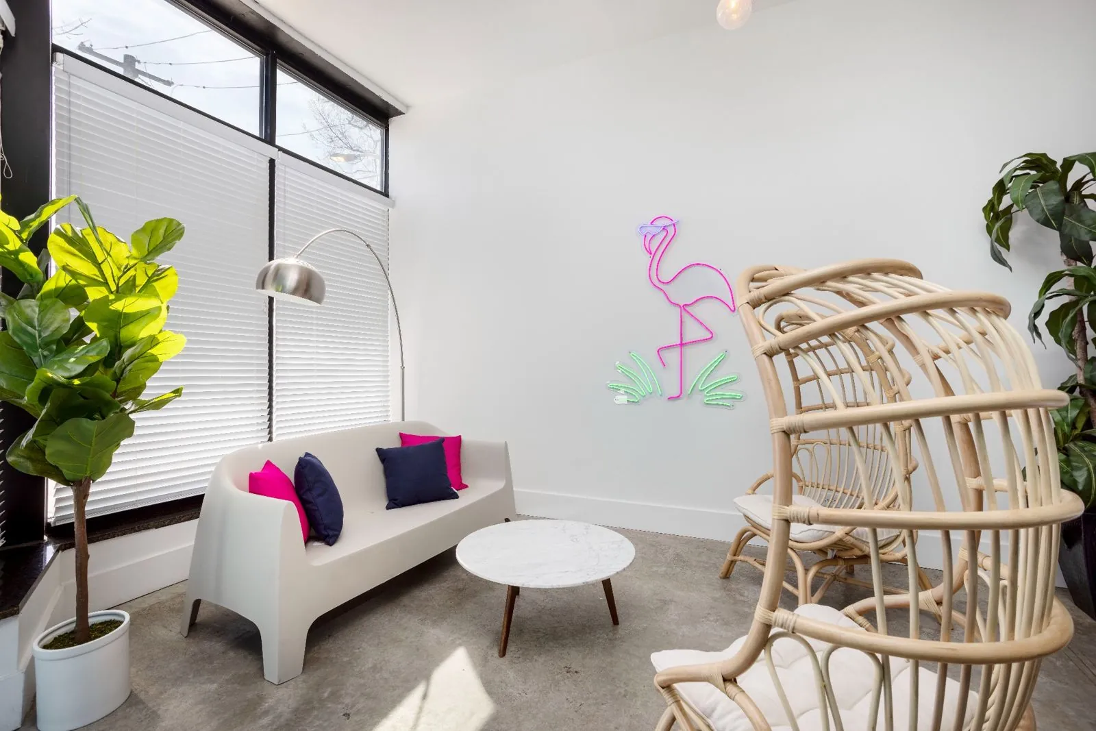 Lounge corner with a white sofa, pink and navy pillows, rattan chairs, and a neon flamingo sign