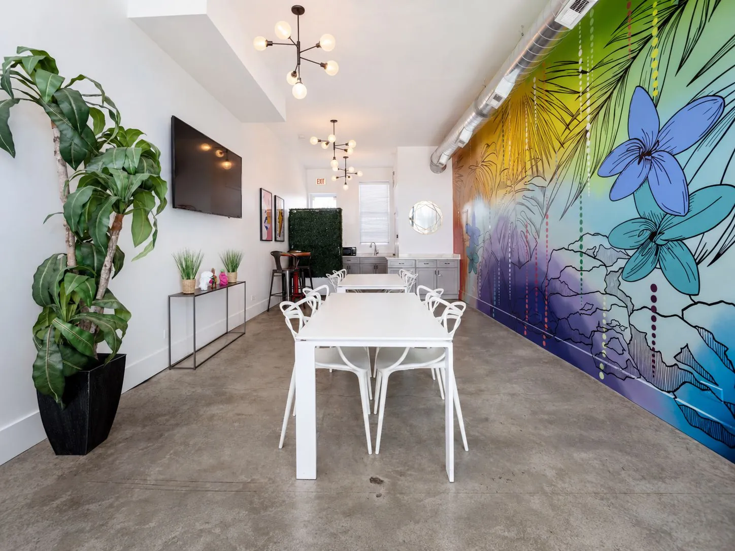 Nala’s Party Room main room with white dining tables, wall-mounted TV, and a colorful tropical mural
